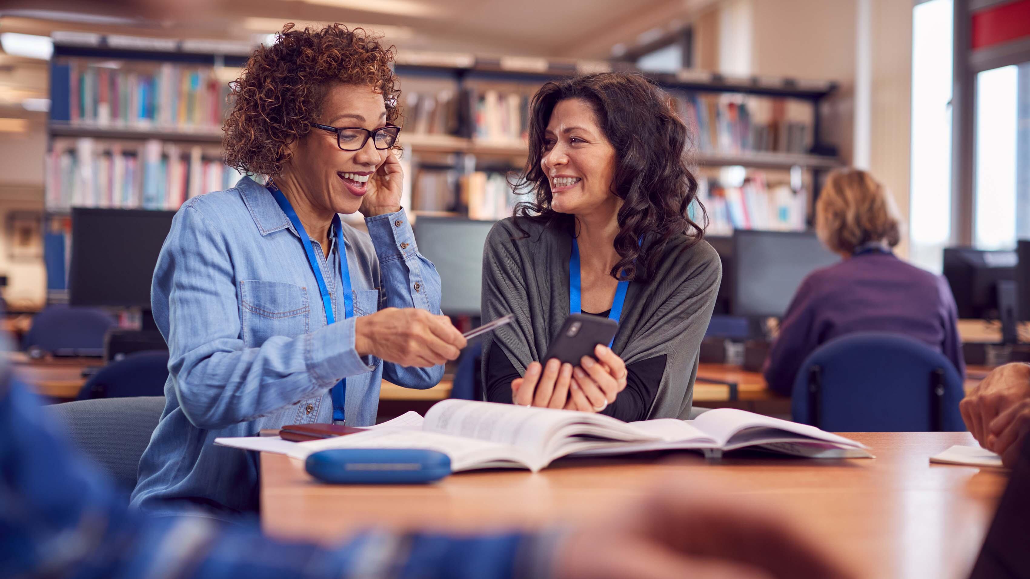 Teacher With Mature Female Adult Student Using Mobile Phone At Table Working In College Library