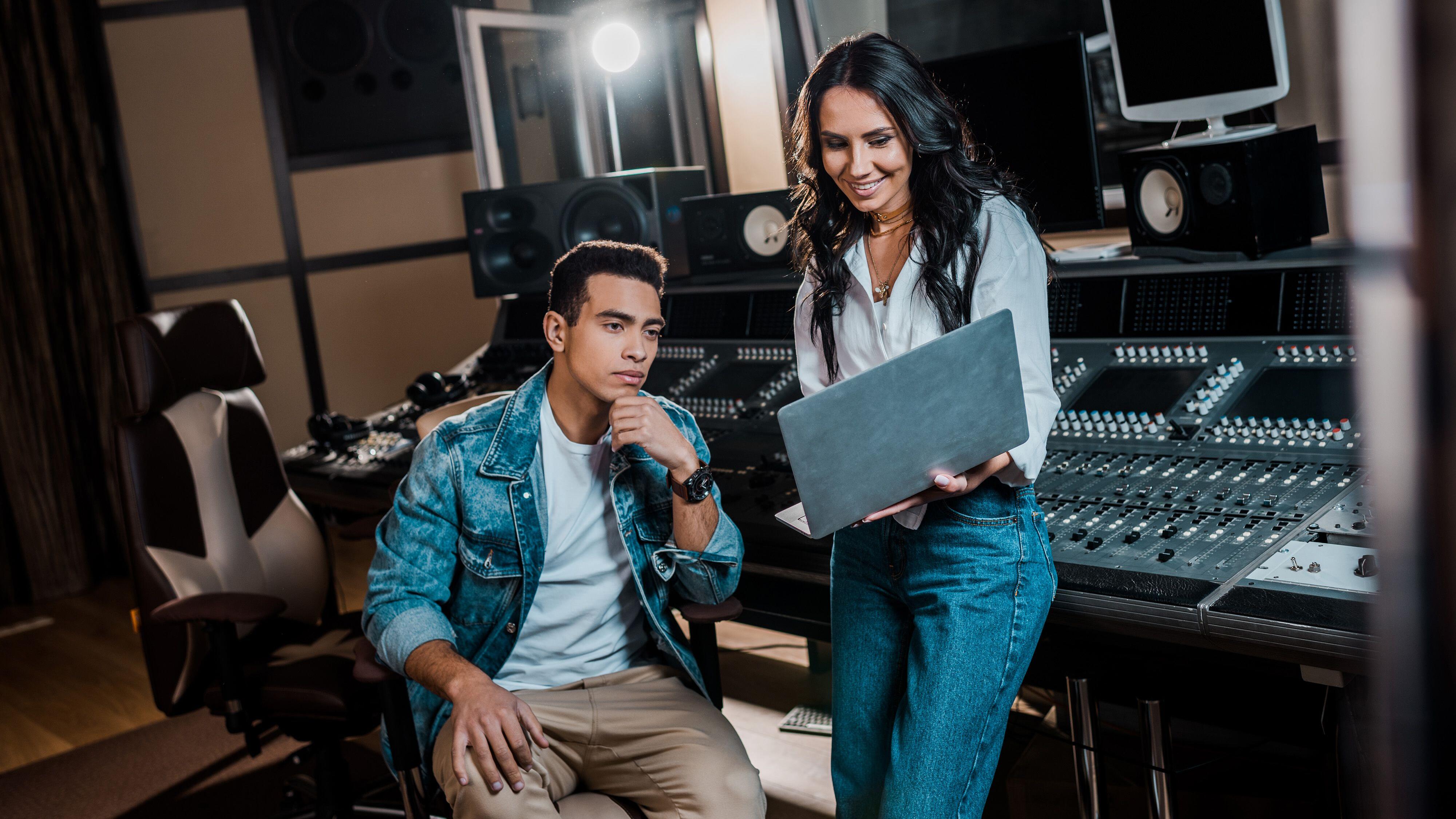 smiling pretty sound producer using laptop near mixed race friend in recording studio