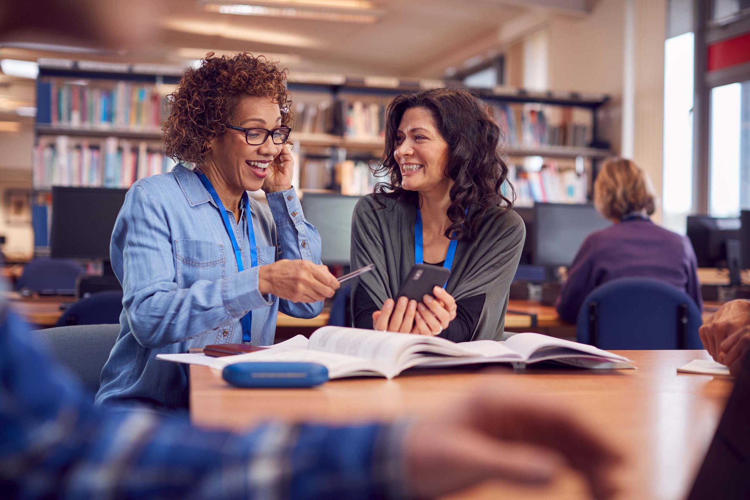 Teacher With Mature Female Adult Student Using Mobile Phone At Table Working In College Library